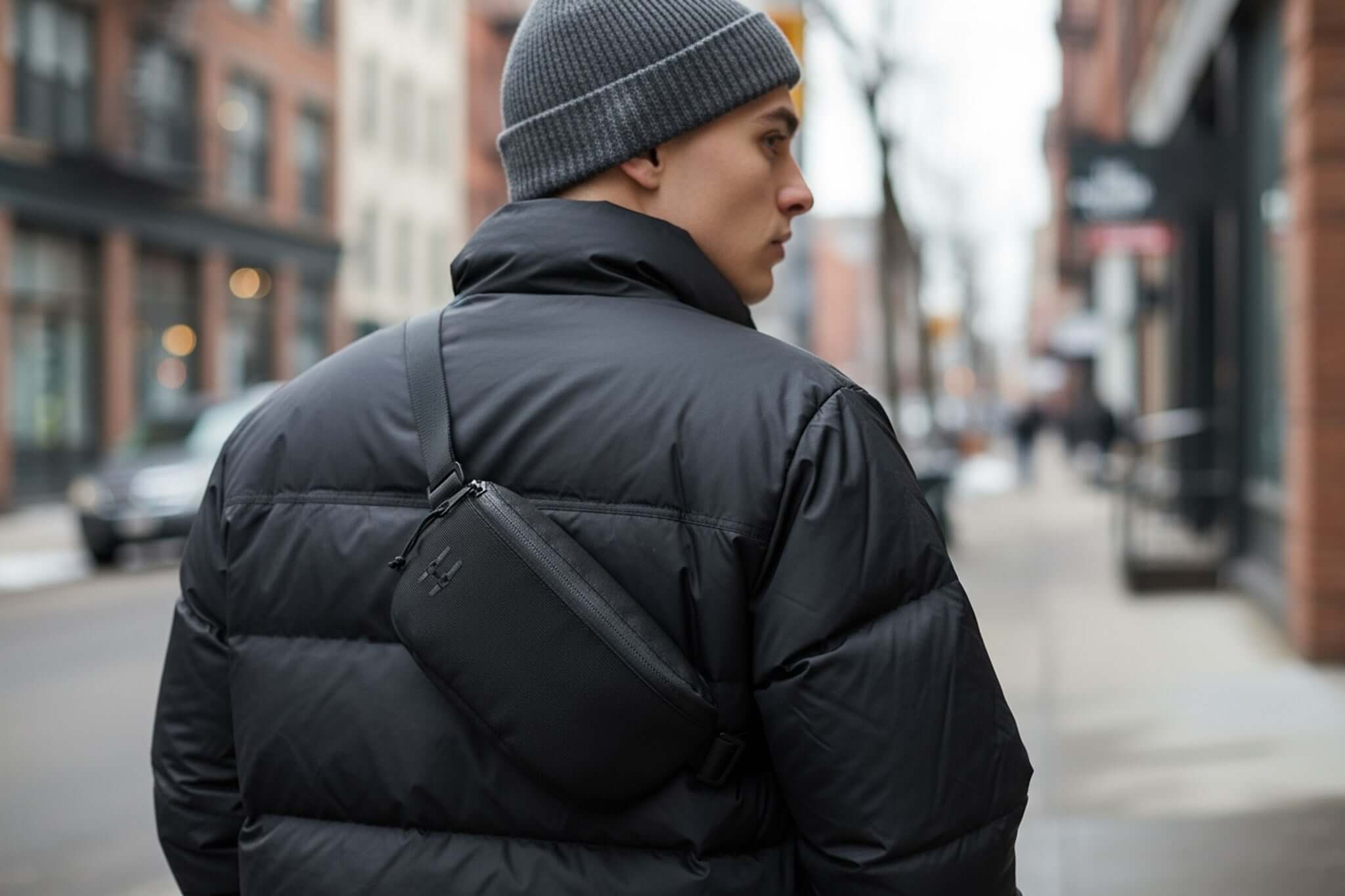 Muž v černé bundě nese ledvinku Sora 2.0 v lesklé černé barvě na ulici v městském prostředí.