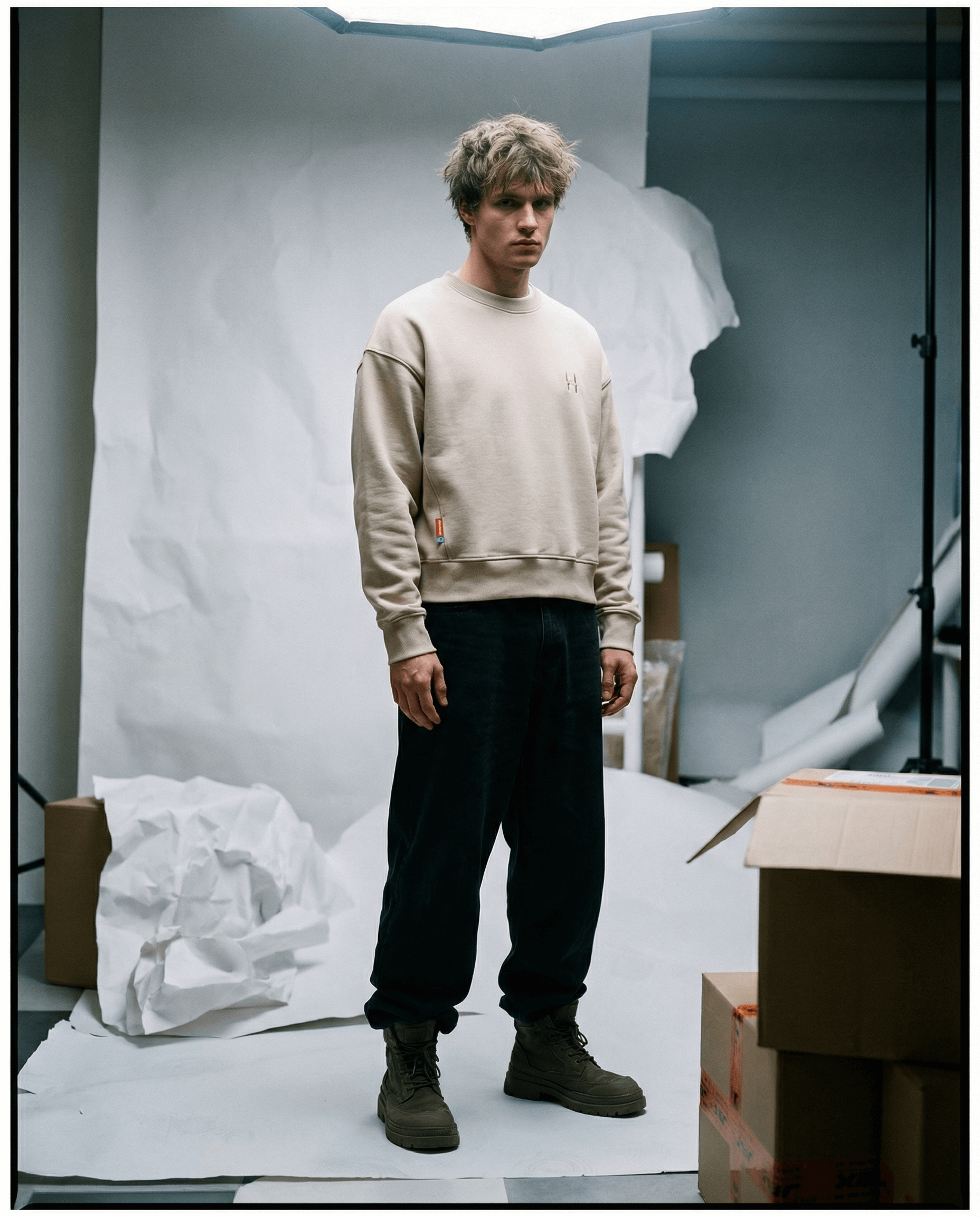 Muž v béžové mikině crewneck s cropped oversized střihem, stojící mezi balíky a papírovými pozadími.
